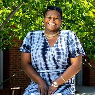 Image of Black female presenting person wearing a blue tie-dye shirt sitting in front a tree with green leaves.